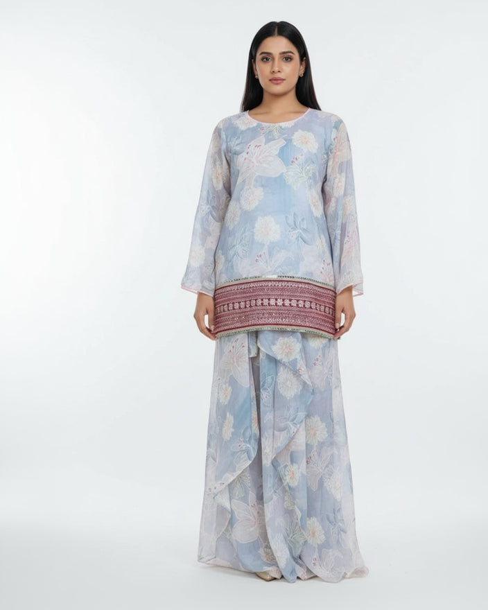 Woman wearing a light blue floral saree with a traditional border in a home setting.