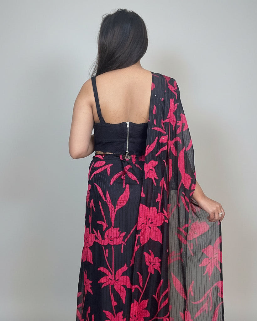 Woman wearing a black and pink floral saree in a room with decorative plates on the wall.