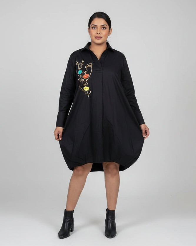 Woman wearing a black dress with colorful design standing in a room with beige walls and tiled floor.