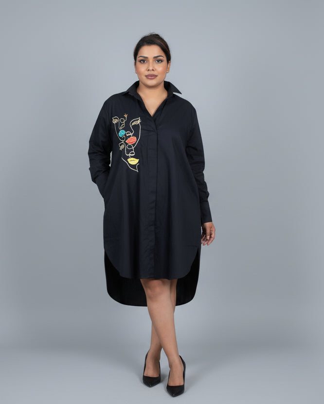 Woman wearing a black dress with colorful embroidery in a room with a white wall and tiled floor.